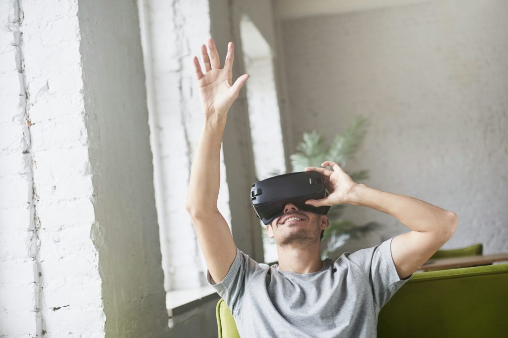 Un jeune homme caucasien attrape quelque chose dans l'air, joue à un casque de réalité virtuelle innovant pour les applications de jeux mobiles, assis sur un canapé à la maison.
