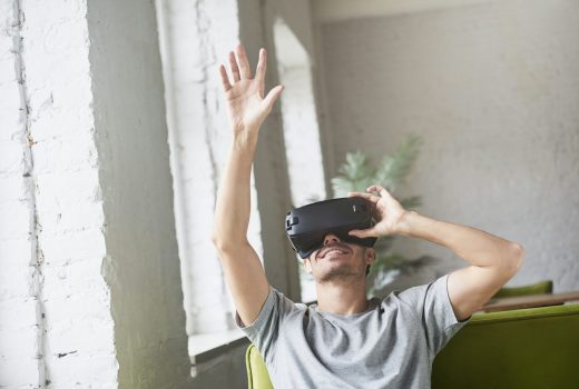 Un jeune homme caucasien attrape quelque chose dans l'air, joue à un casque de réalité virtuelle innovant pour les applications de jeux mobiles, assis sur un canapé à la maison.