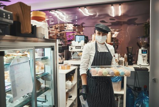 Dame responsable en uniforme de chef portant un masque et tenant un récipient rempli de macarons tout en posant dans un café confortable