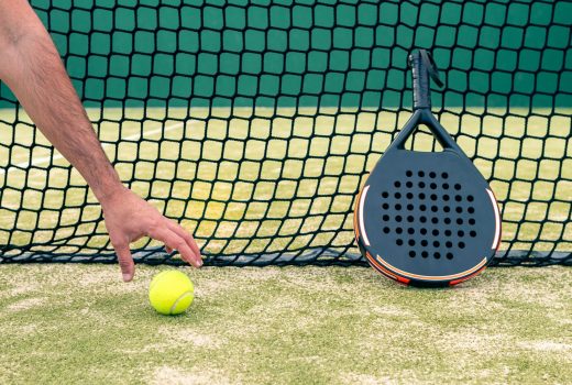 Un homme de race blanche attrape une balle jaune près de la raquette de padel sur un gazon vert en plein air au coucher du soleil.