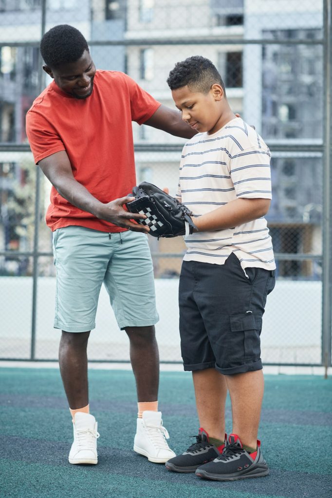 Père afro-américain positif en t-shirt rouge montrant comment attraper la balle tout en expliquant les règles du baseball à son fils