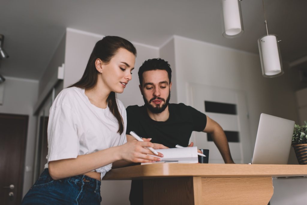 Angle bas de couple sérieux dans des vêtements décontractés assis à une table en bois avec un ordinateur portable et en prenant des notes tout en travaillant ensemble